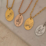 Four engraved necklaces with hand gesture designs, including peace signs and hand-holding, displayed on a marble background.