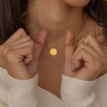 Close-up of a gold pendant necklace with an engraved outline of a US state, worn by a woman holding it up with two hands.