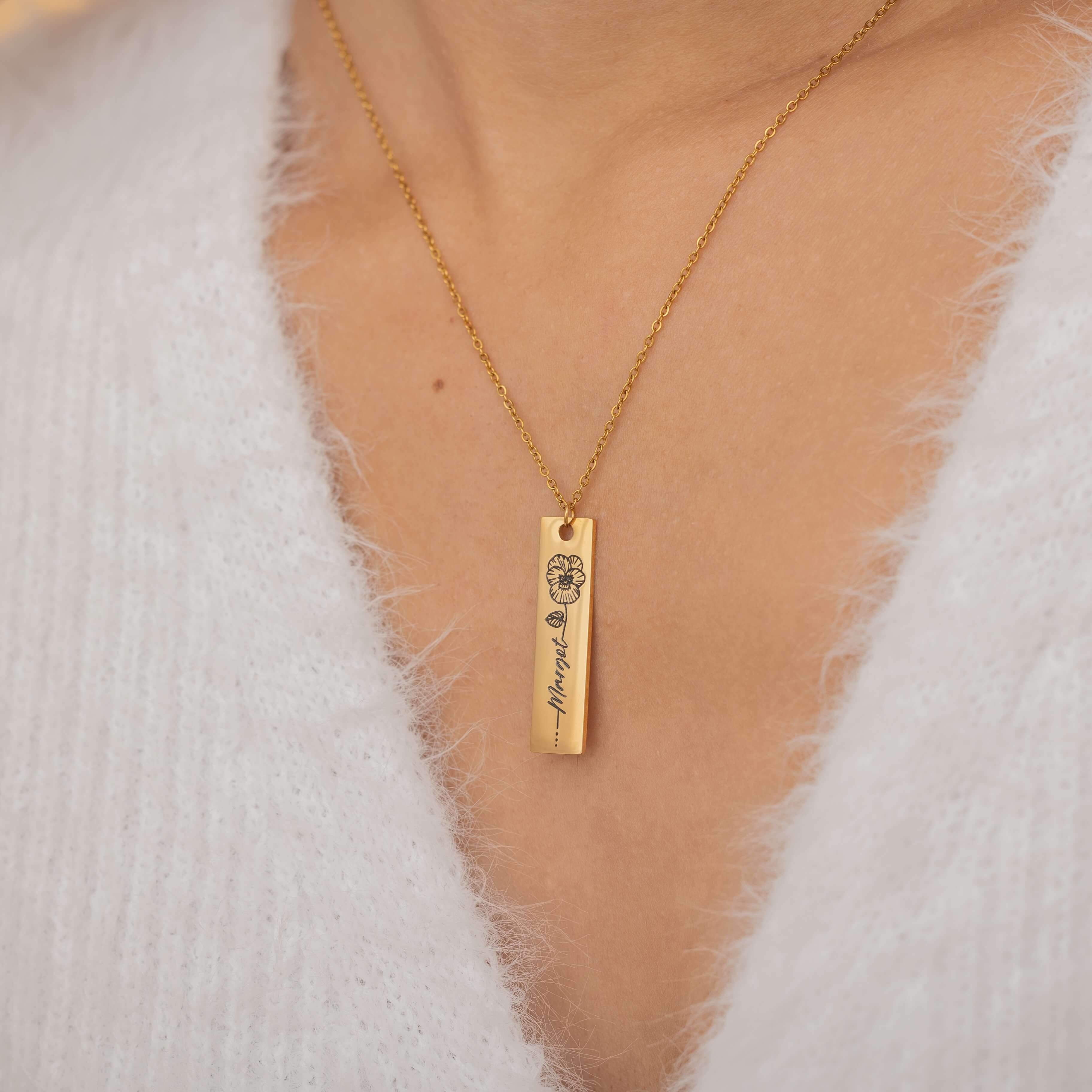Close-up of a gold pendant necklace featuring an engraved birth flower with a personalized name as the stem, worn by a woman.