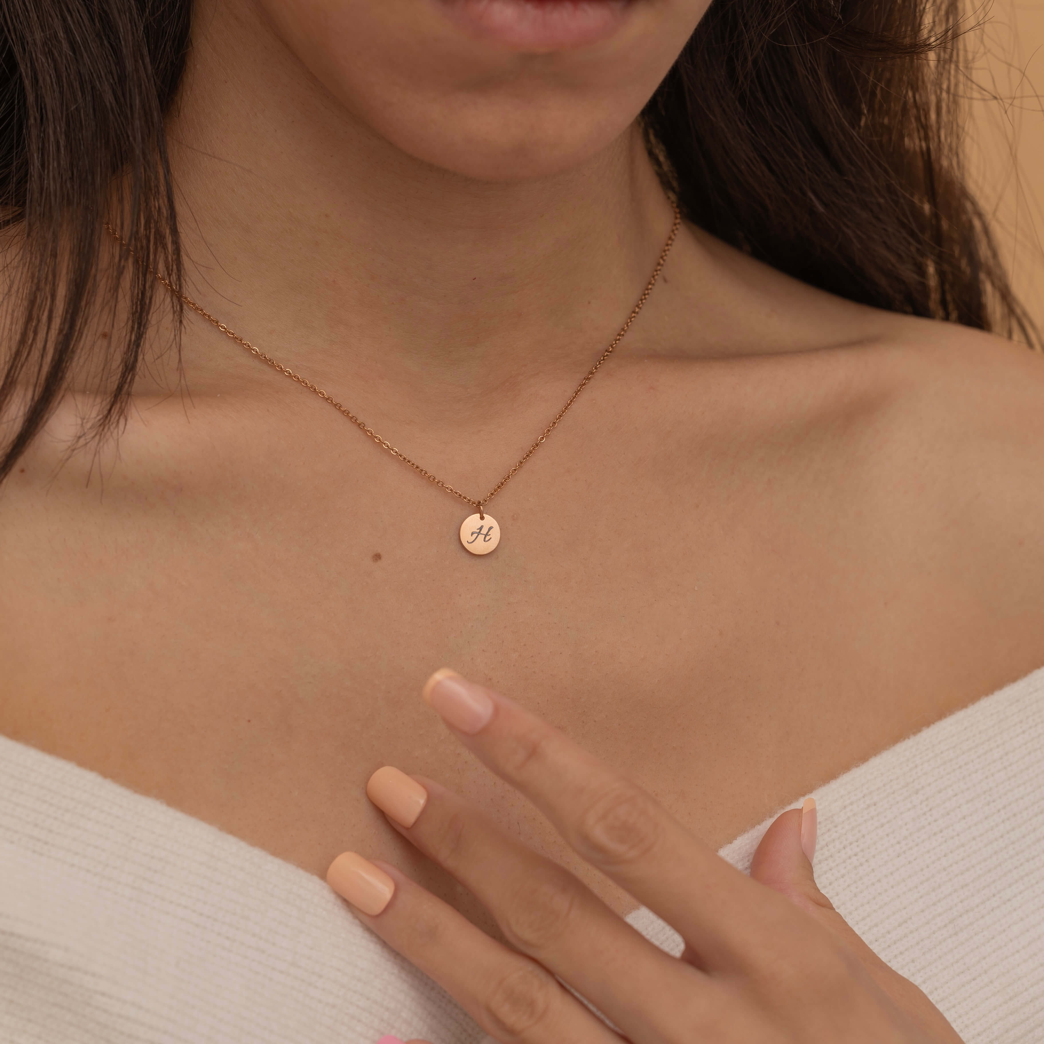 Close-up of a rose gold pendant necklace with an engraved initial "H" worn by a woman touching her chest.