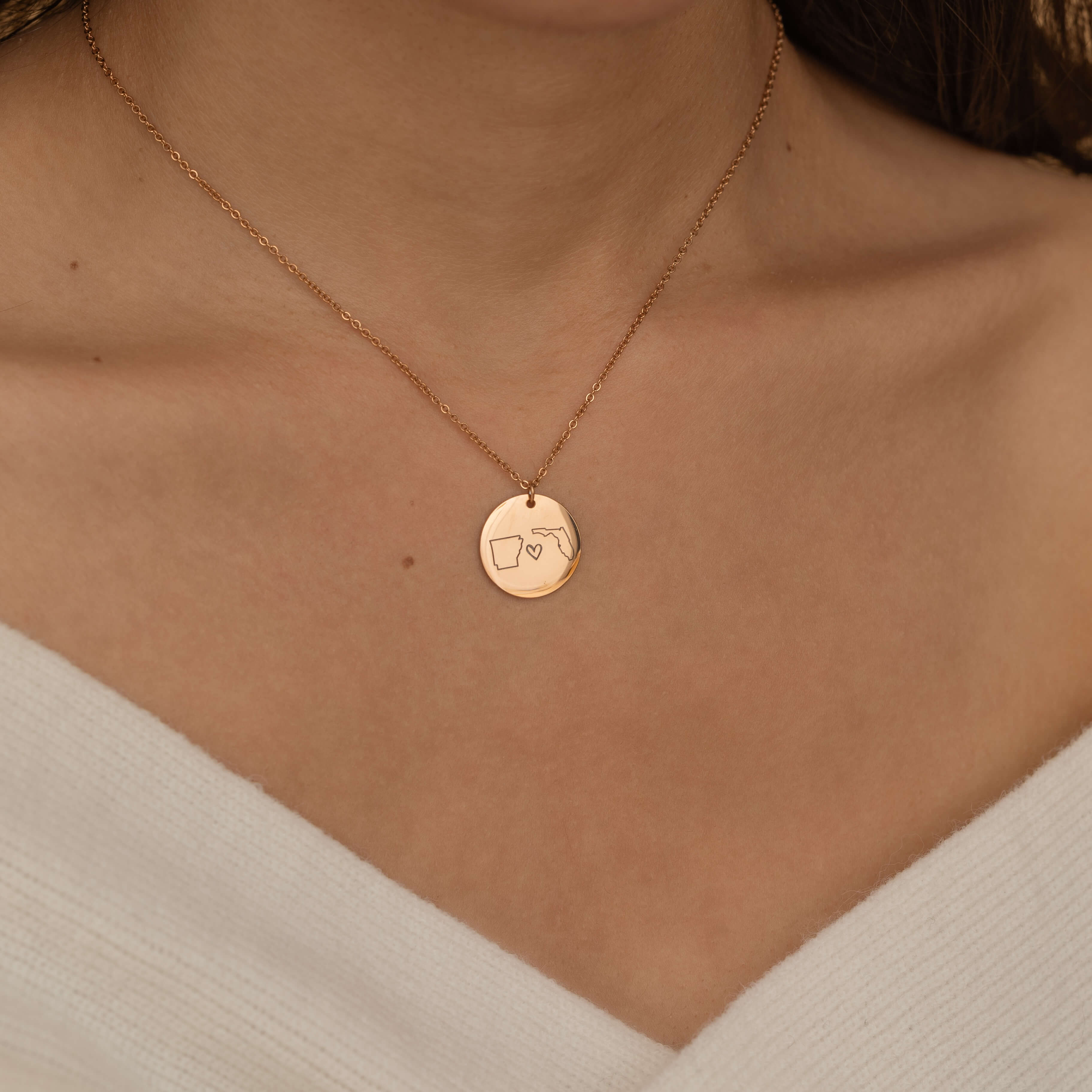 Close-up of a rose gold pendant necklace engraved with two state outlines and a heart in the center, worn by a woman.