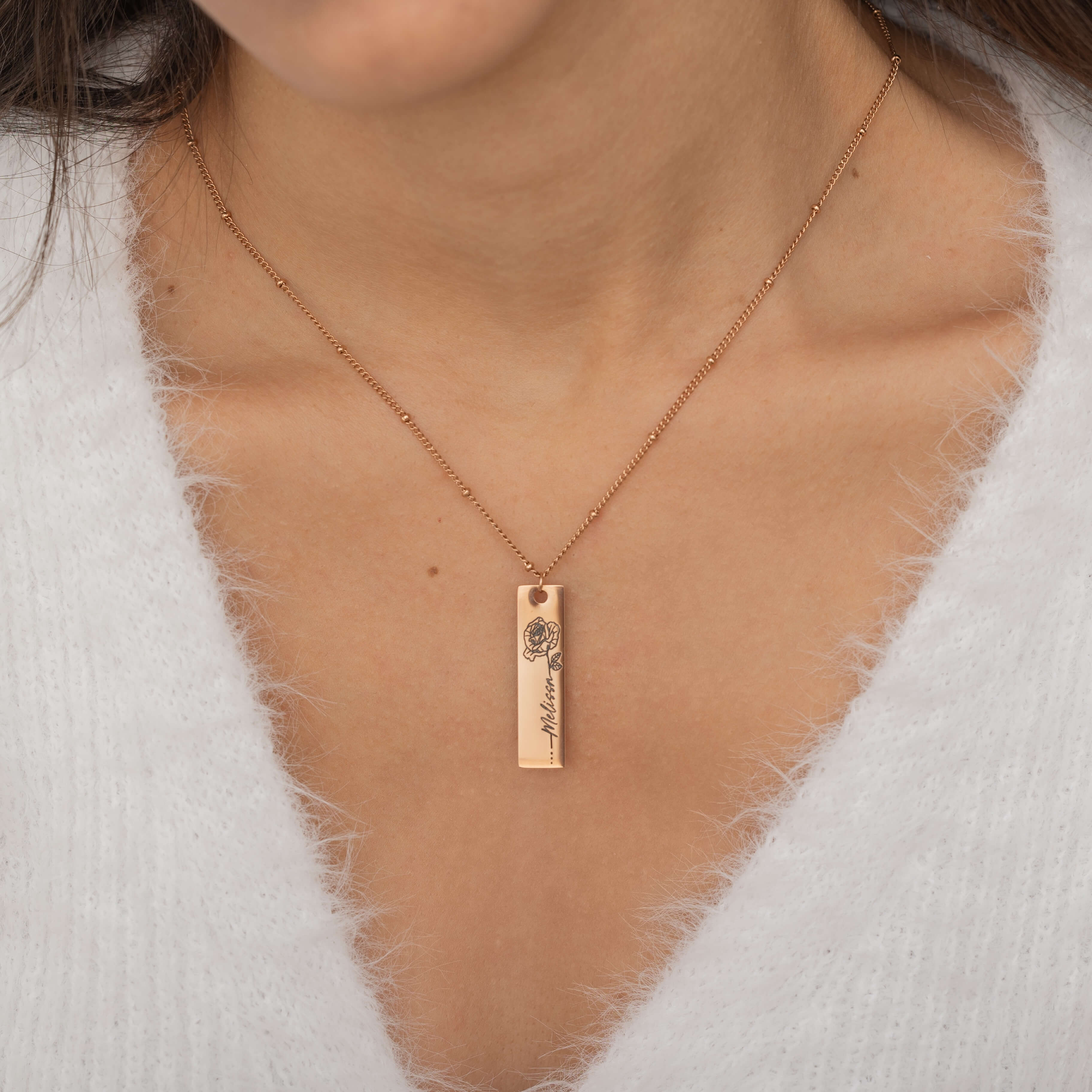 Close-up of a rose gold pendant necklace featuring an engraved birth flower with a personalized name as the stem, worn by a woman.