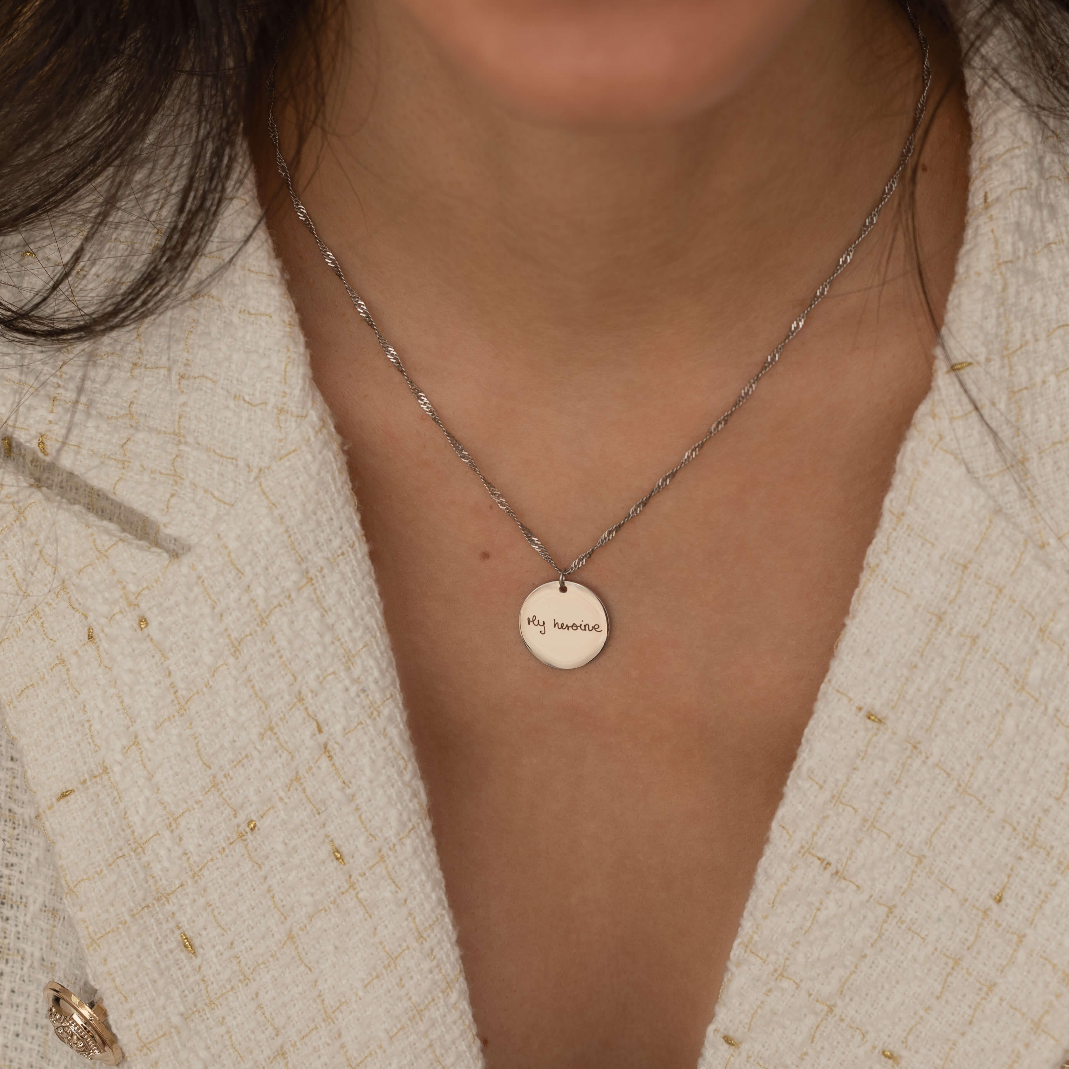 Close-up of a silver round pendant necklace engraved with the handwritten message "My heroine", worn by a woman.