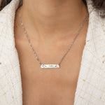 Close-up of a silver bar necklace engraved with two state outlines at each corner and a heart in the middle connecting them, worn by a woman.