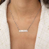 Close-up of a silver bar necklace engraved with two state outlines at each corner and a heart in the middle connecting them, worn by a woman.