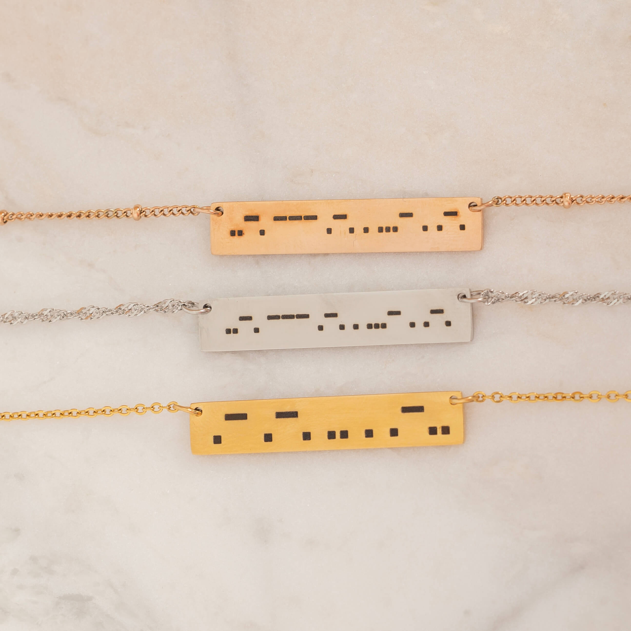 Close-up of three bar necklaces each engraved with a personalized Morse code message, on top of a stone.