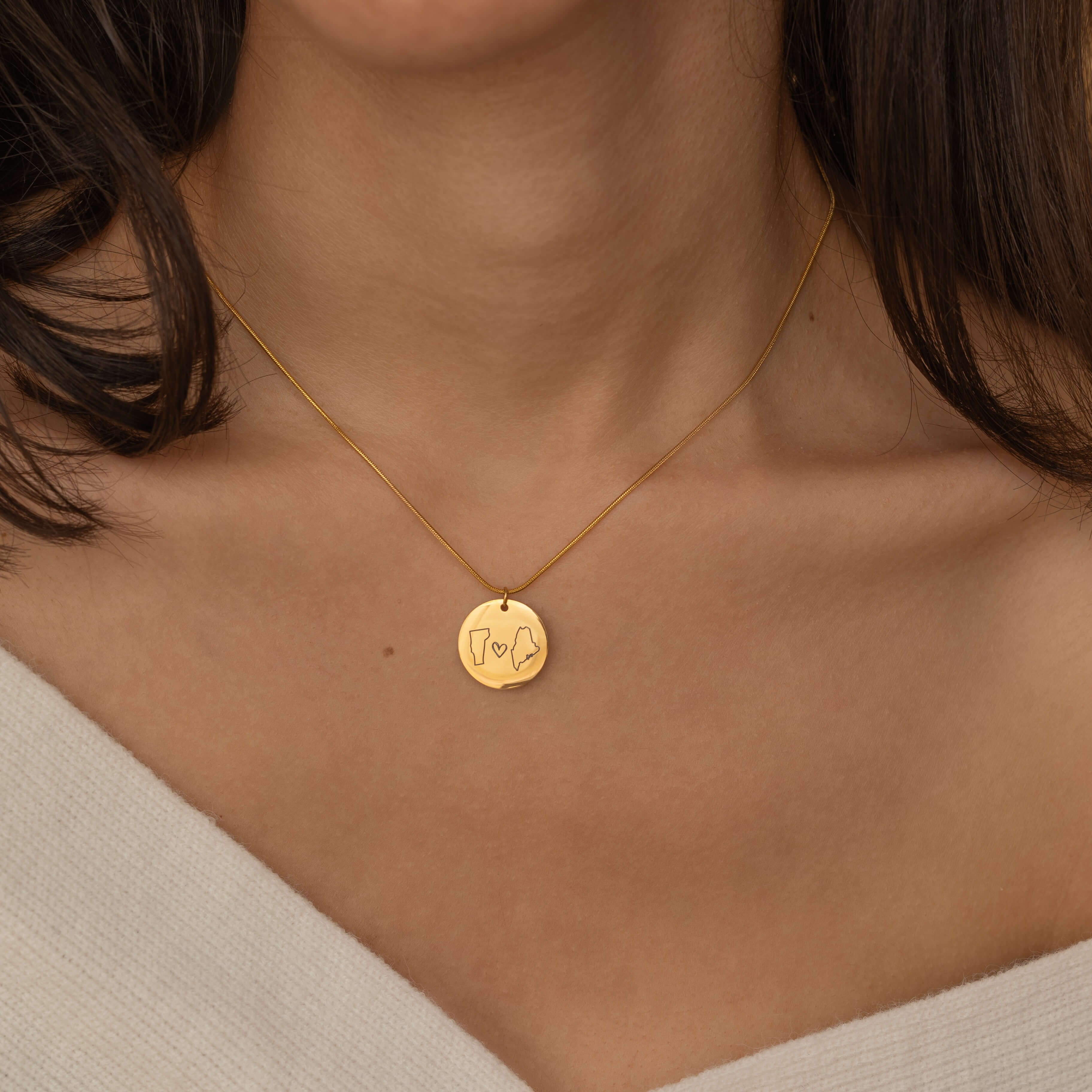 Close-up of a gold pendant necklace engraved with two state outlines and a heart in the center, worn by a woman.
