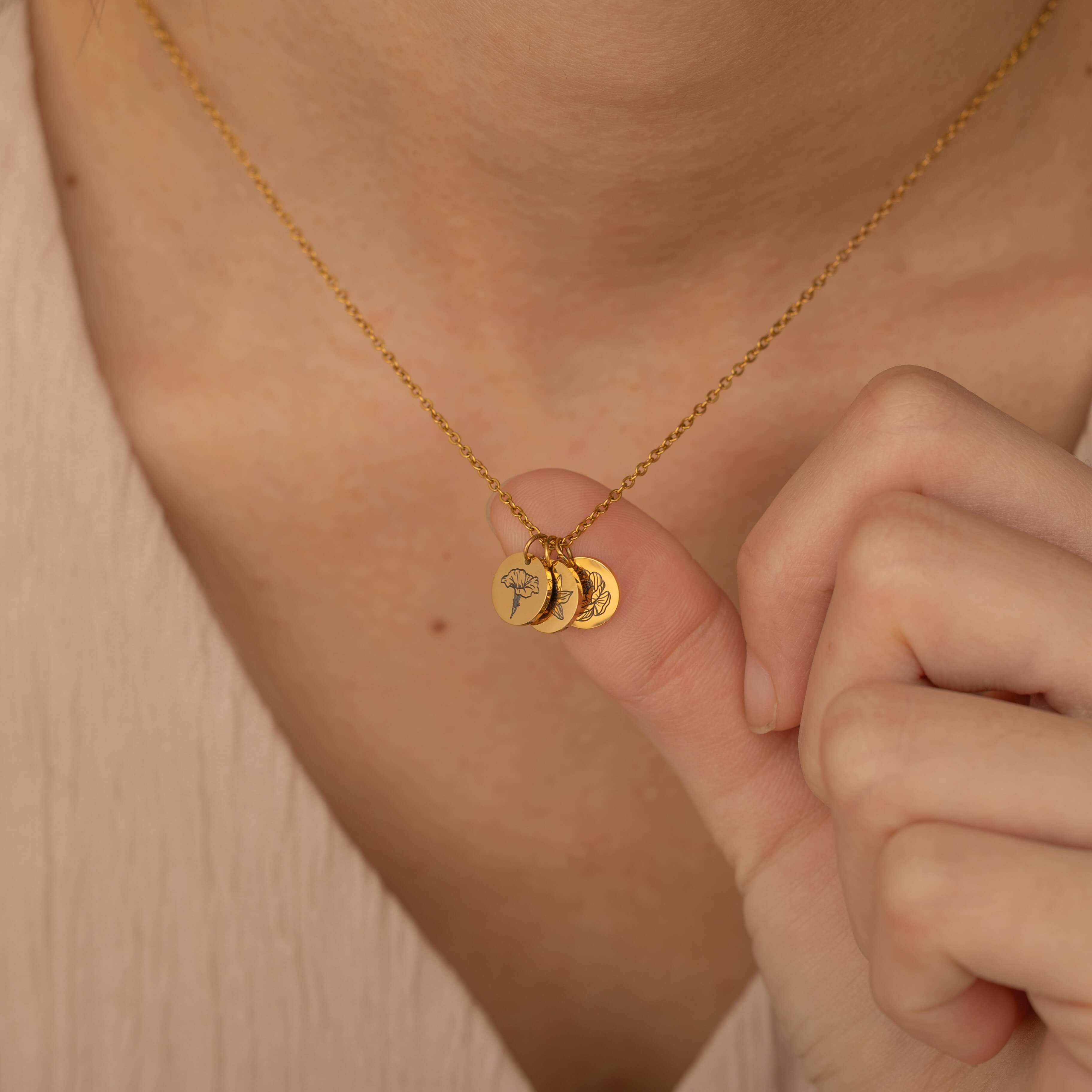 Close-up of a gold custom birth flower engraved pendant necklace worn by a woman, showcasing three round charms with floral engravings being held between fingers.