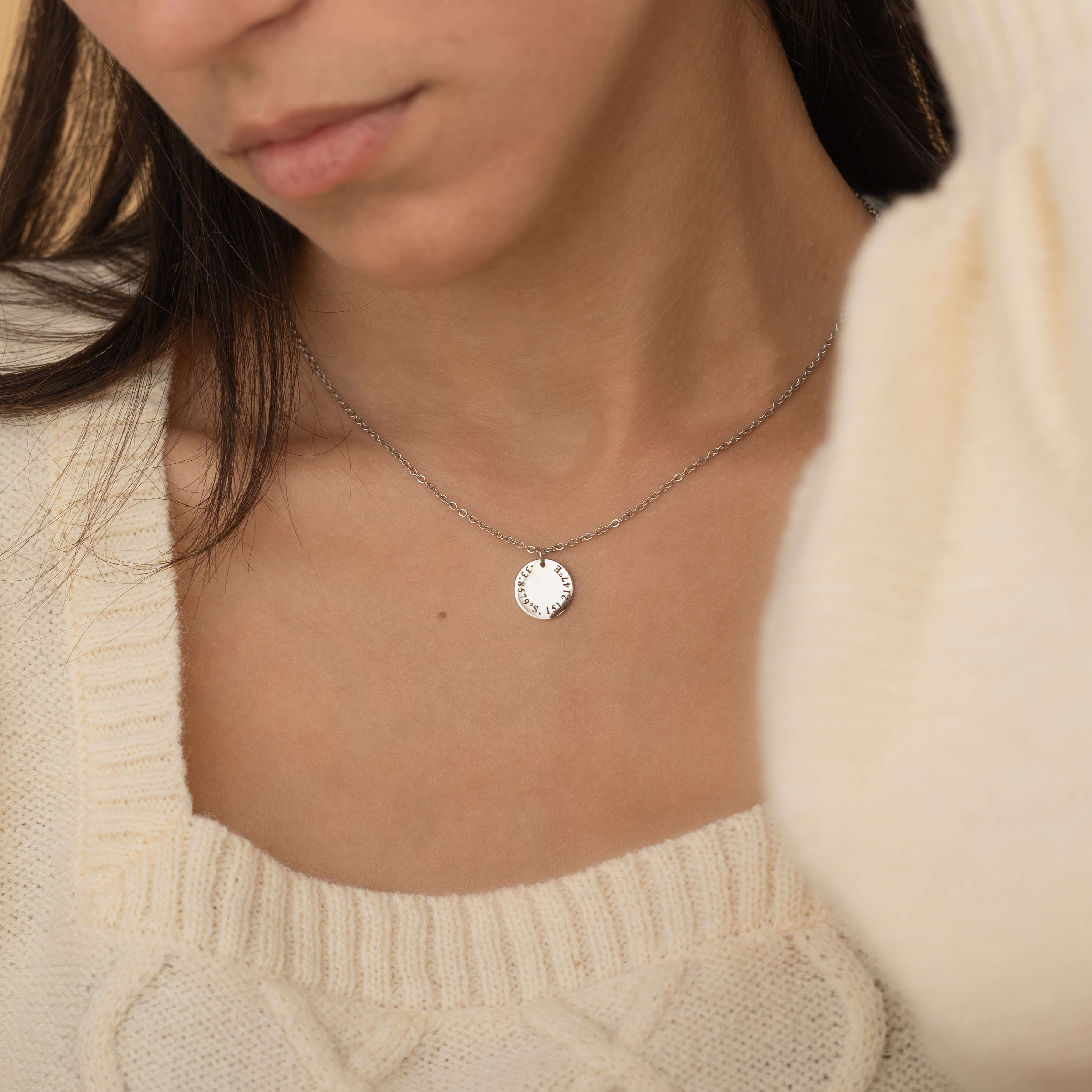 Close-up of a silver custom coordinate necklace worn on a woman's neck, featuring engraved latitude and longitude details on a round pendant.