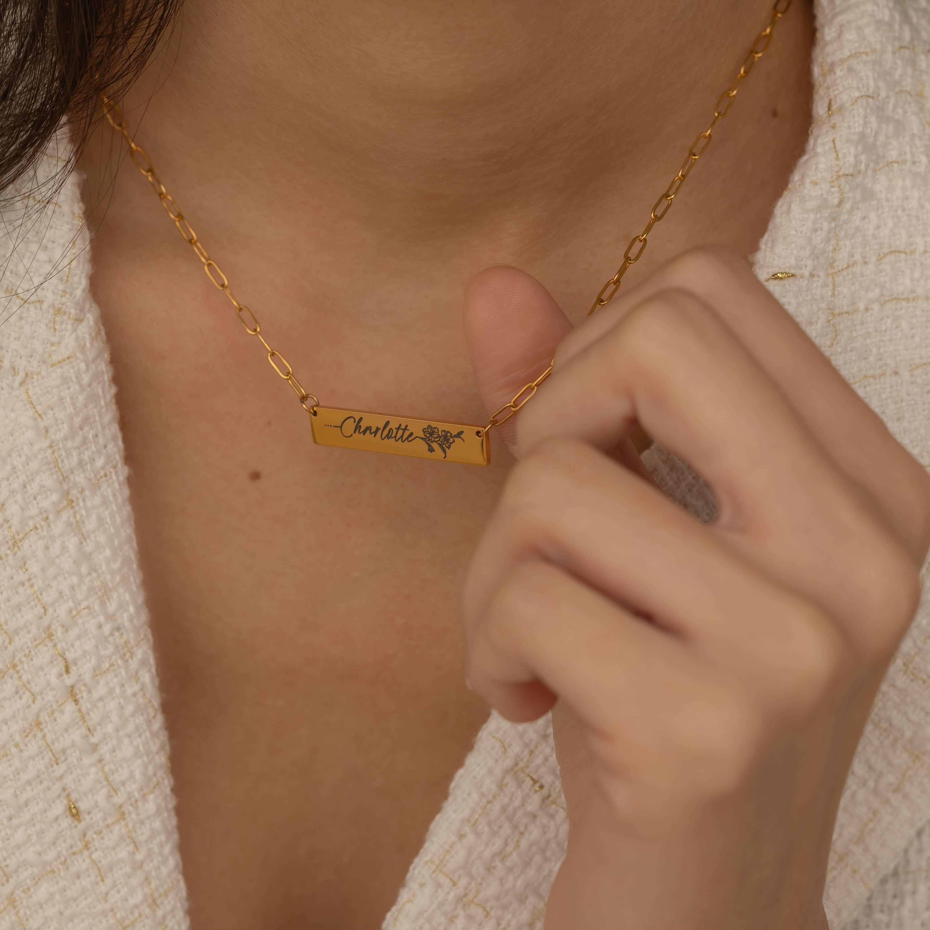 Close-up of a gold bar necklace with a personalized birth flower design and a name engraved as the flower's stem, worn by a woman.