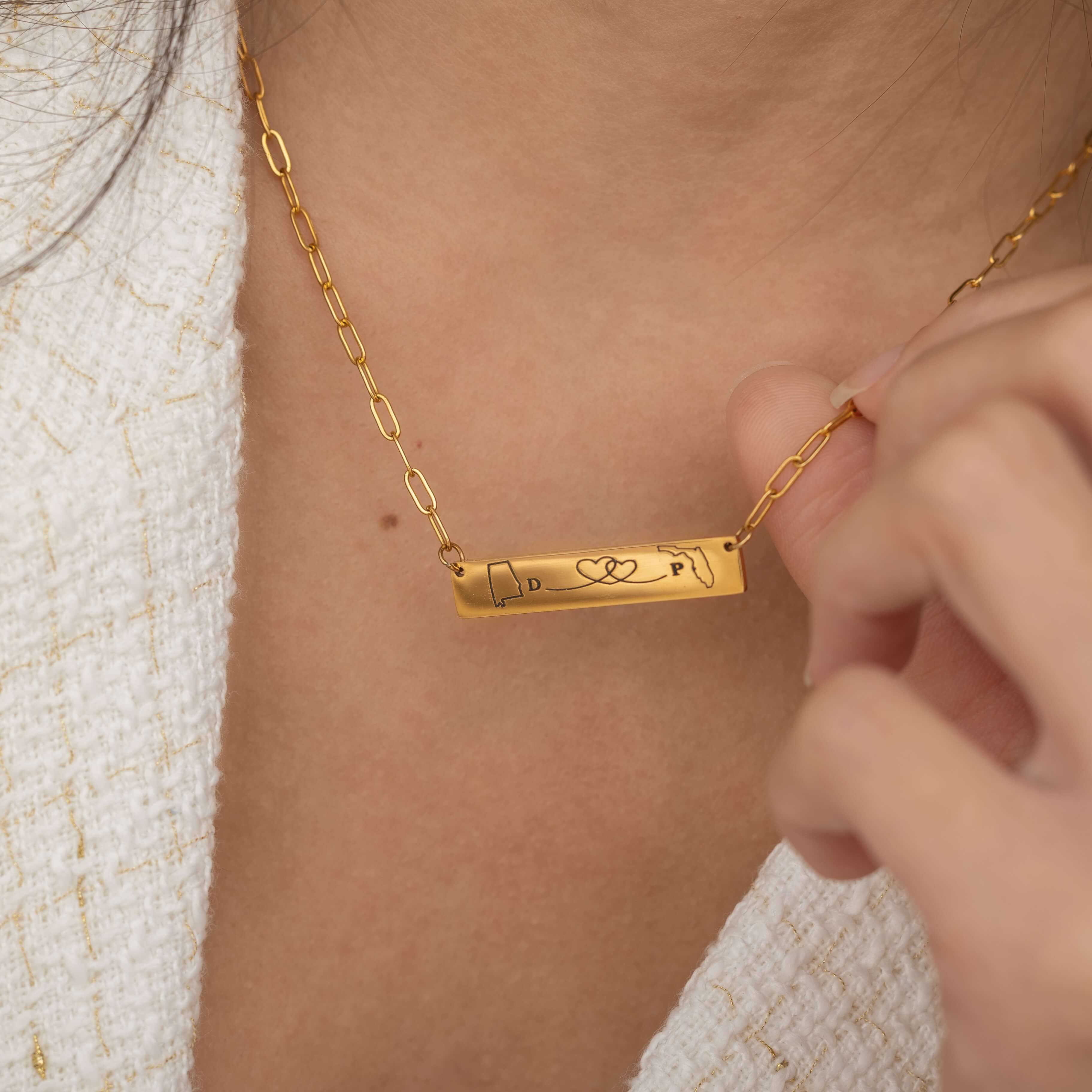 Close-up of a gold bar necklace engraved with two state outlines at each corner and a heart in the middle connecting them, worn by a woman.
