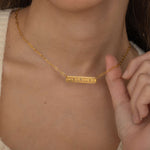 Close-up of a gold bar necklace engraved with three state outlines and two connecting hearts between them, worn by a woman.