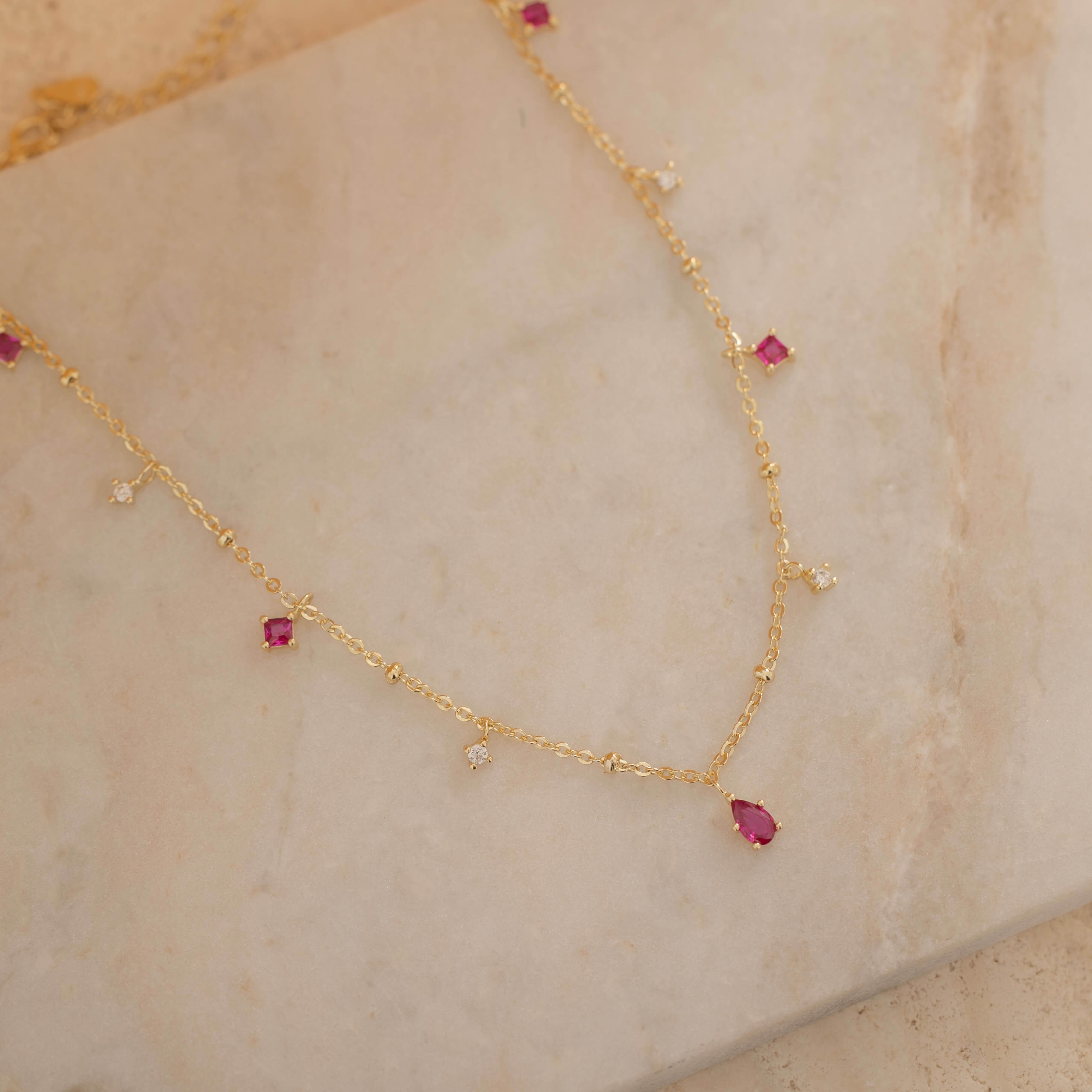 Dainty sterling silver and 14k gold plated necklace featuring four square pink ruby-colored cubic zirconia stones, one centered teardrop-shaped pink ruby stone, and four white cubic zirconia accents between the pink ruby. 