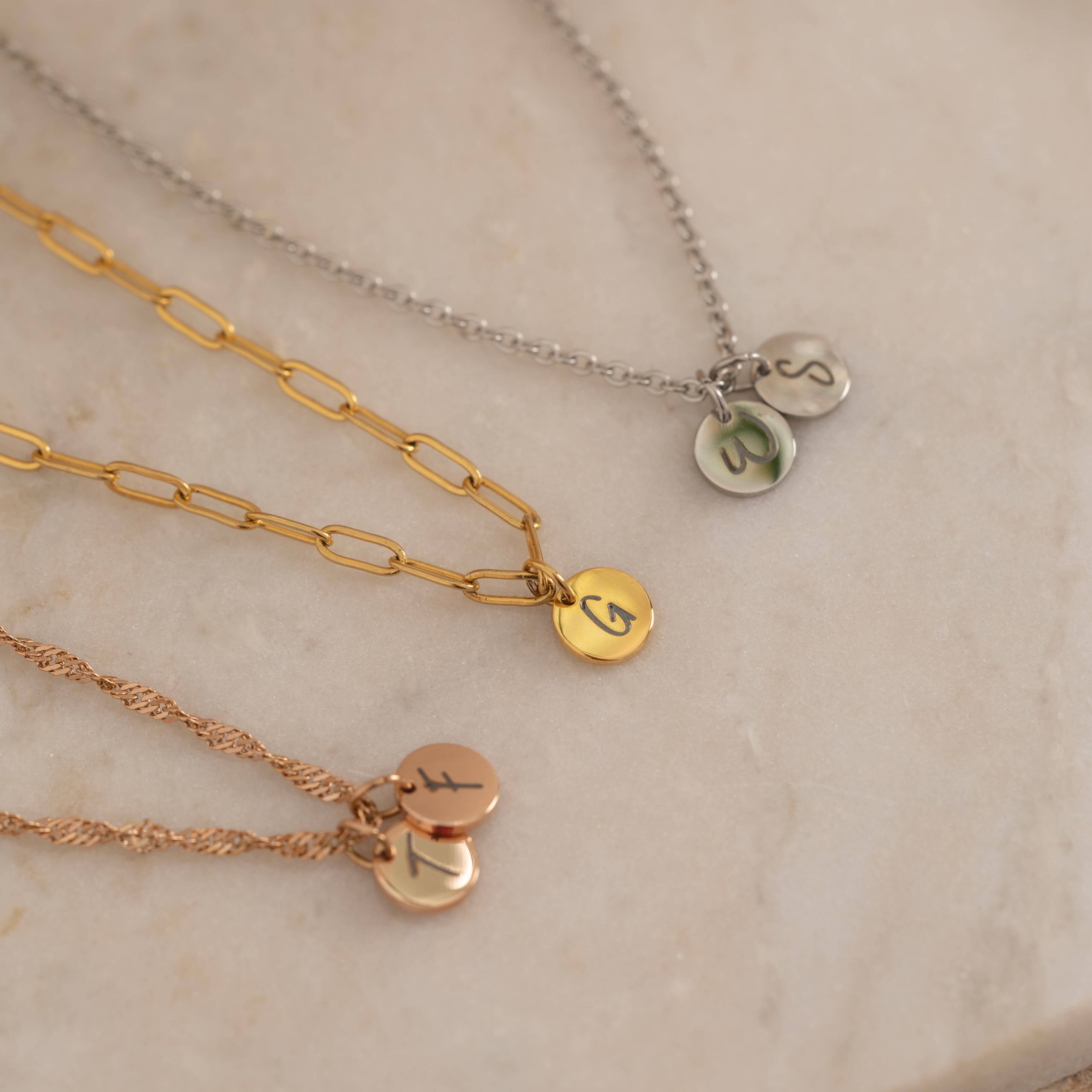 Three custom initial charm necklaces in gold, silver, and rose gold finishes, each with distinct chain styles and personalized round charms on a marble surface.