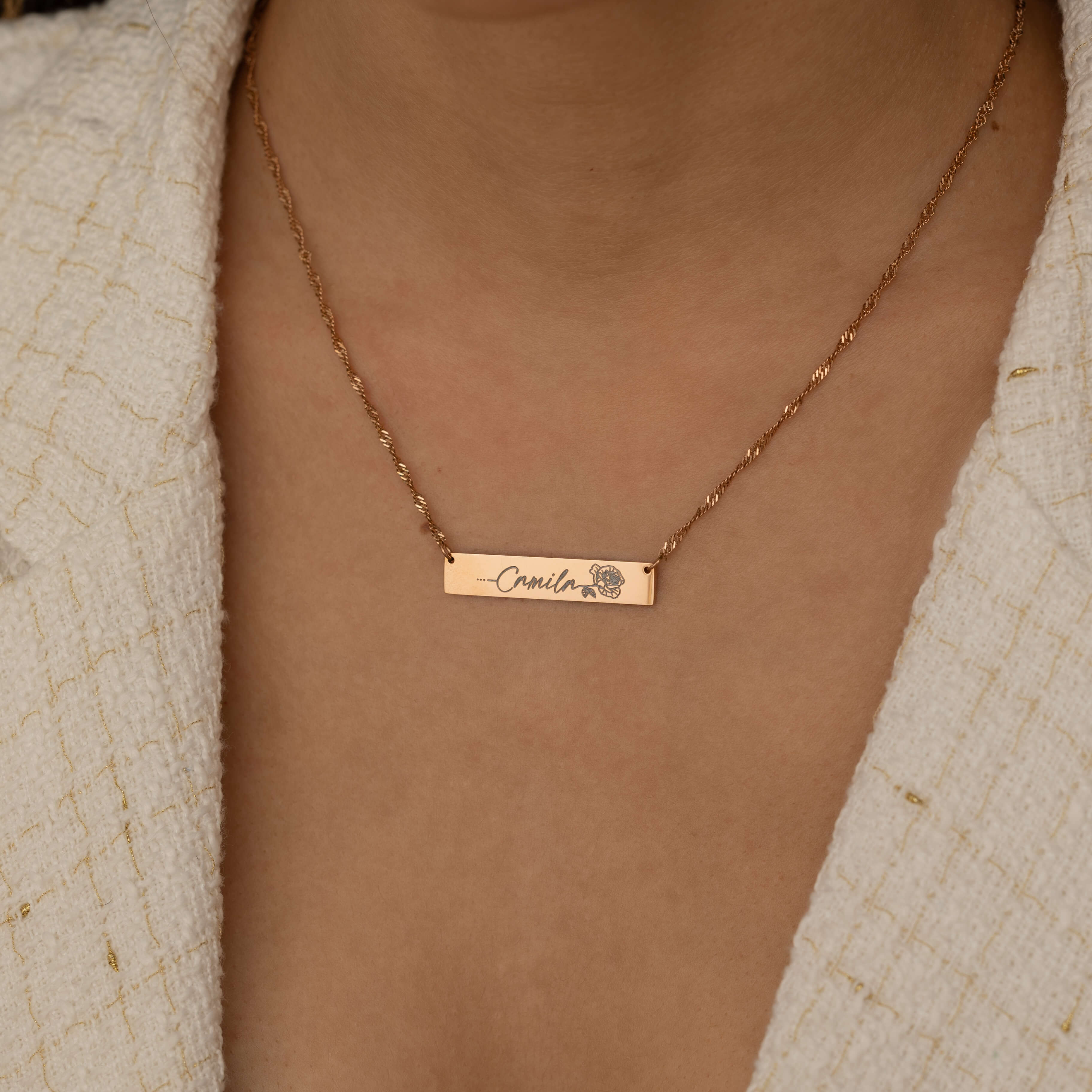 Close-up of a rose gold bar necklace with a personalized birth flower design and a name engraved as the flower's stem, worn by a woman.