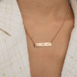Close-up of a rose gold bar necklace engraved with two state outlines at each corner and a heart in the middle connecting them, worn by a woman.