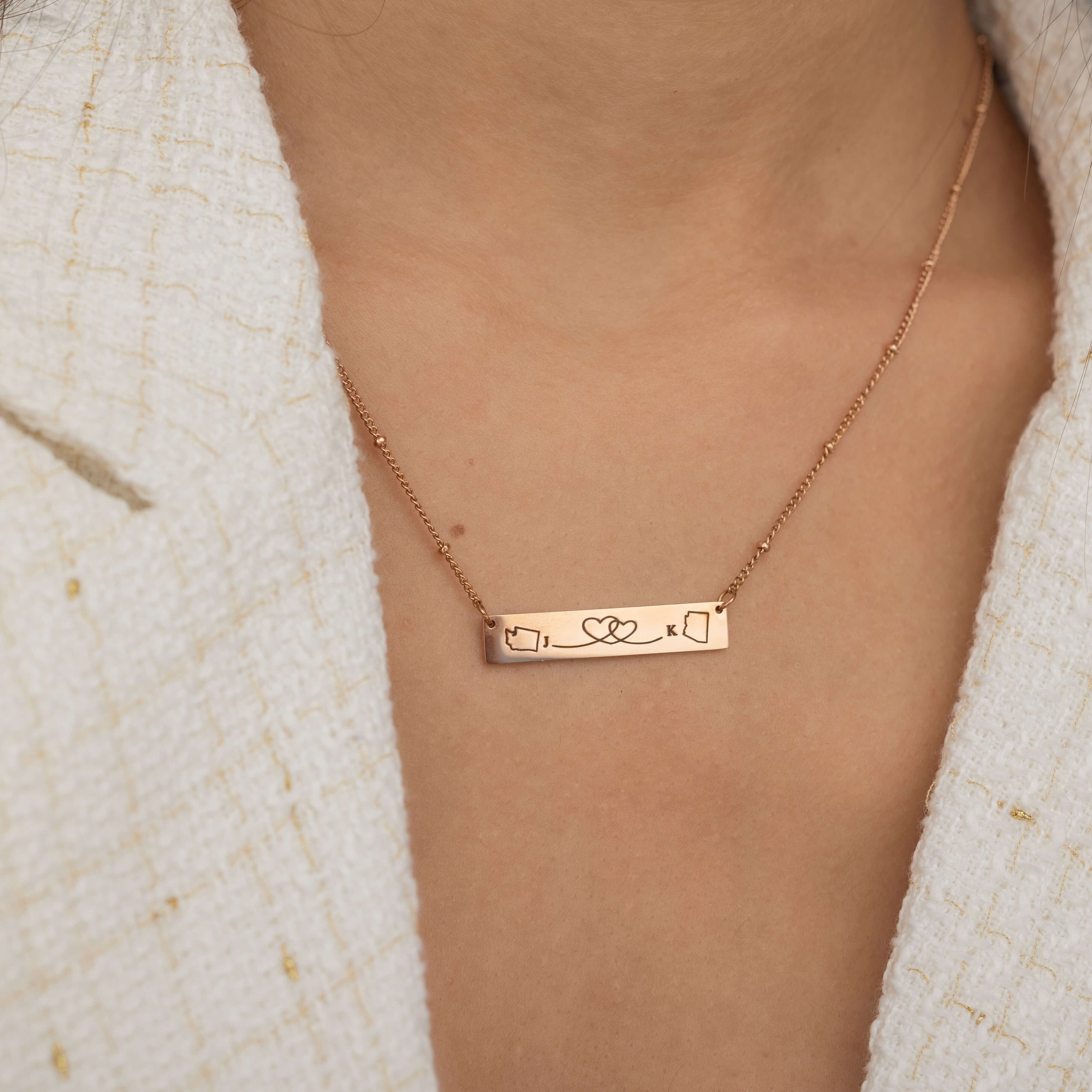 Close-up of a rose gold bar necklace engraved with two state outlines at each corner and a heart in the middle connecting them, worn by a woman.
