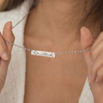 Close-up of a silver bar necklace engraved with two state outlines at each corner and a heart in the middle connecting them, worn by a woman.