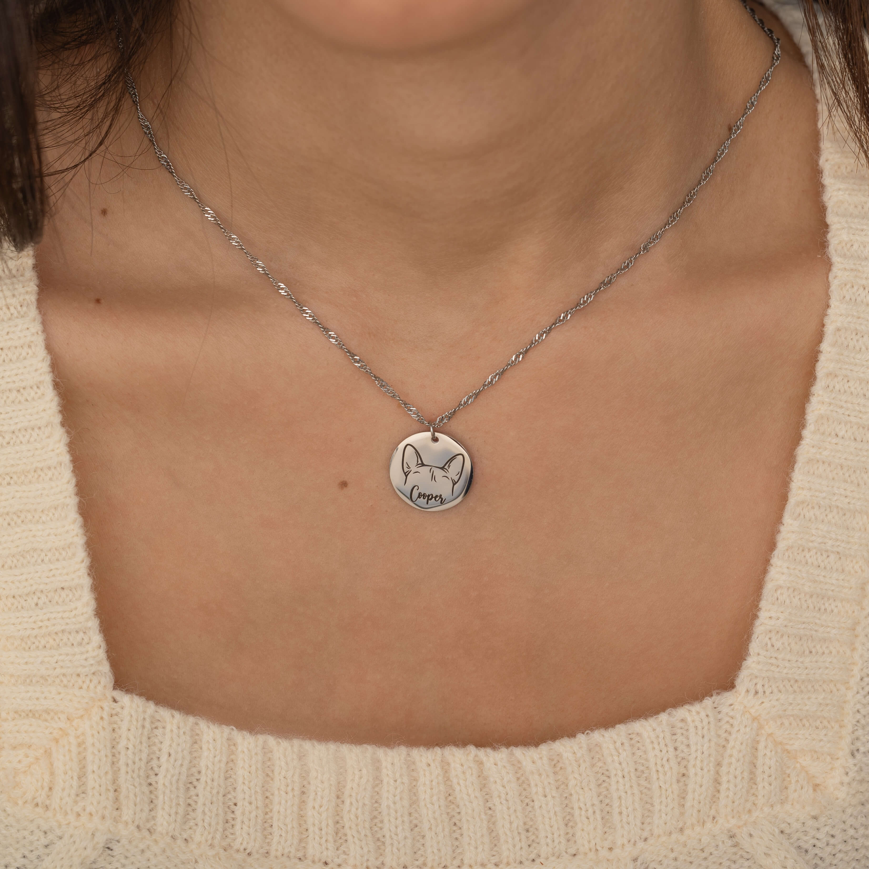 Silver pendant necklace featuring an engraved outline of dog ears with a pet's name underneath, worn by a woman.