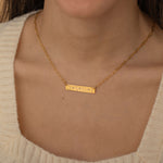 Close-up of a gold bar necklace engraved with three state outlines and two connecting hearts between them, worn by a woman.