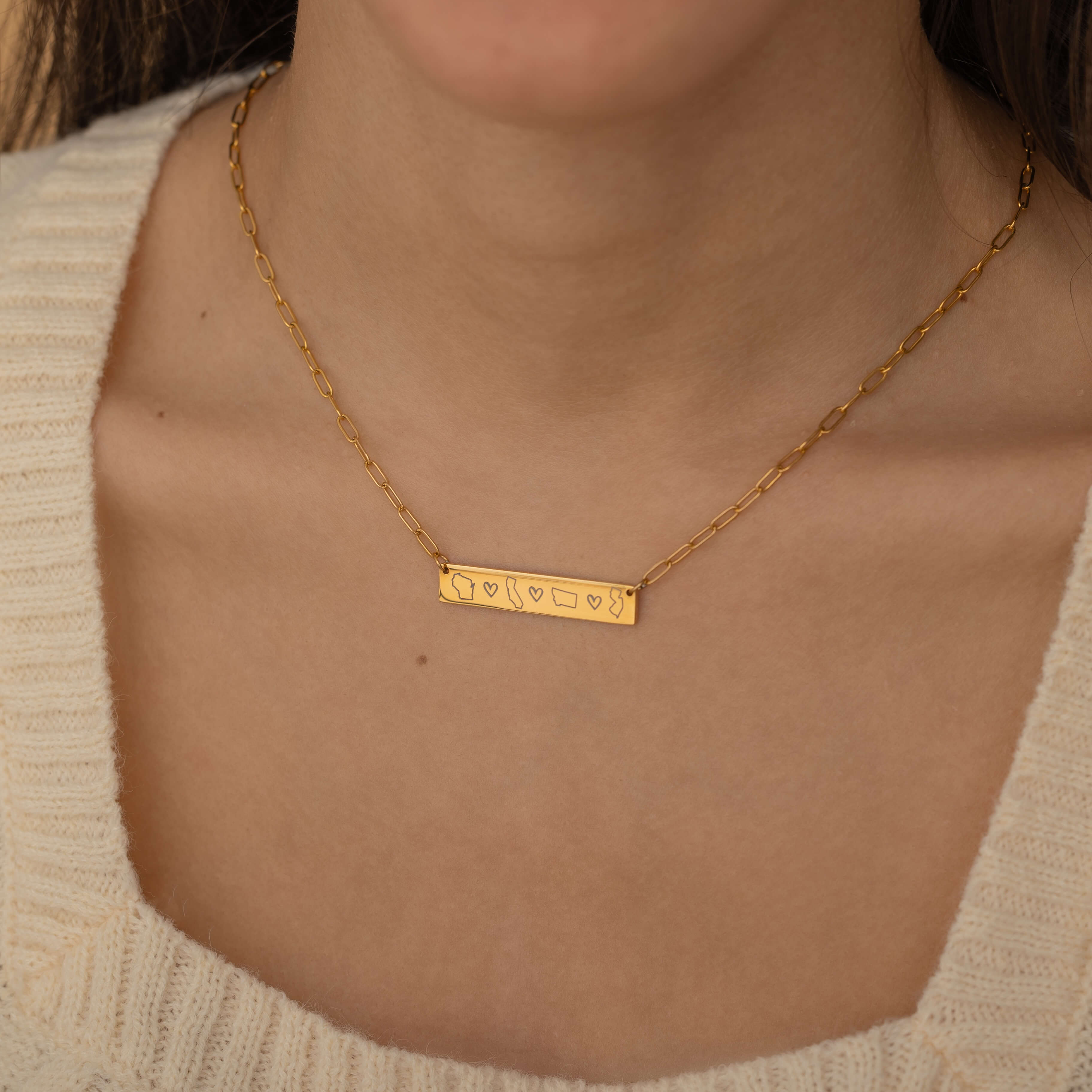 Close-up of a gold bar necklace engraved with three state outlines and two connecting hearts between them, worn by a woman.