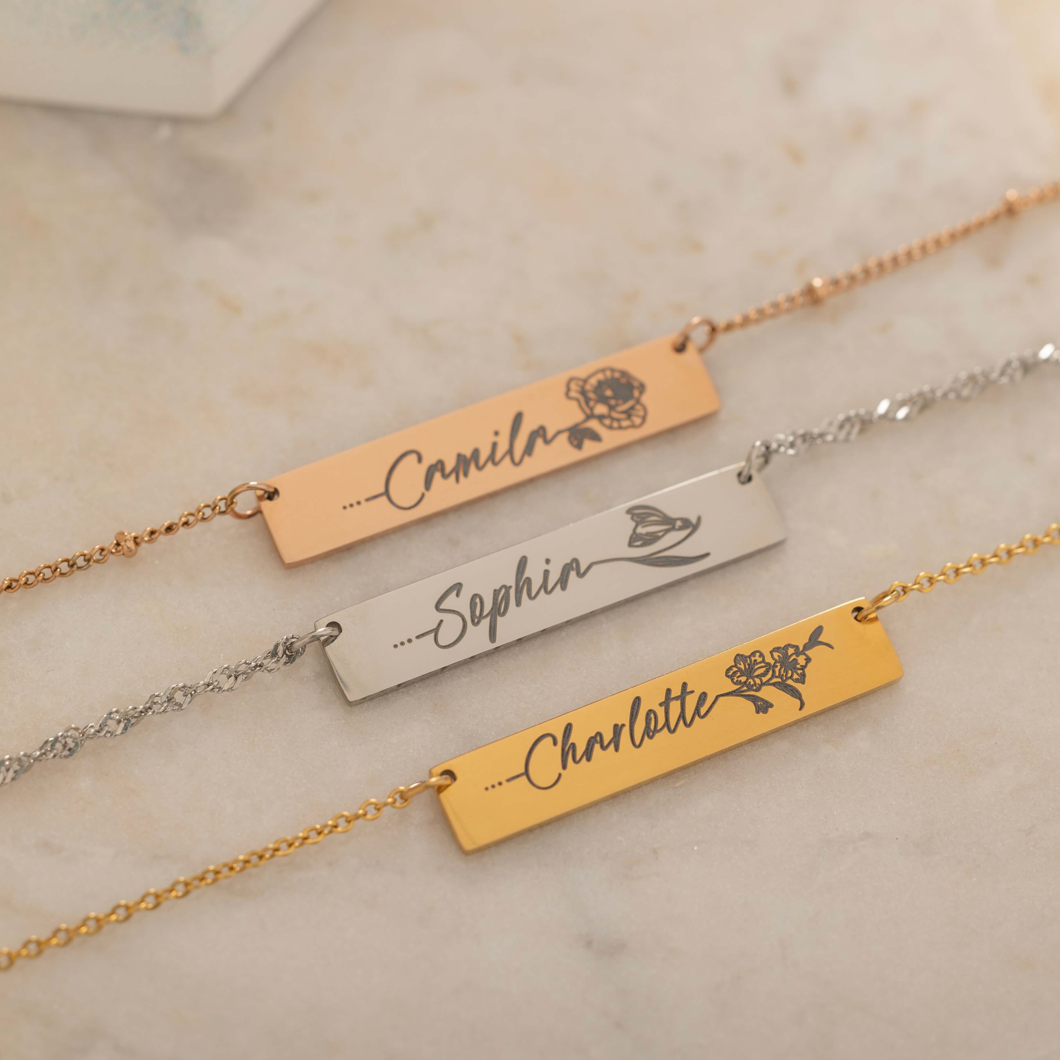 Close-up of three gold bar necklace with a personalized birth flower design each and names engraved as the flower's stem.